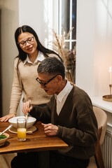 A woman stands with her eyes closed and smiles while placing her hand on the neck of her grandfather who is sitting in front of her at the table and eating, in the kitchen