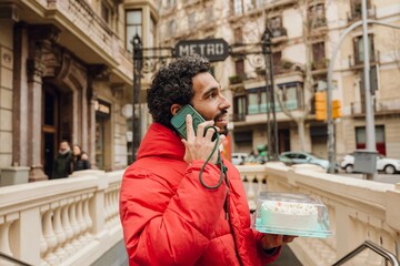 A man stands and holds the phone while talking and holds a cake in his other hand and looks in front of him, on the street near the subway entrance, in the city, preparing for a birthday party