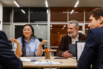 A man sits at a table and looks at a woman who, together with a man, sits next to him and looks at a man sitting next to them, they smile, in the office, at a meeting
