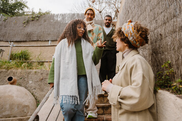 A multiethnic group of four people, dressed warmly is engaged in conversation outdoors near rustic architecture, discussing something while walking down a wooden staircase in a natural setting.