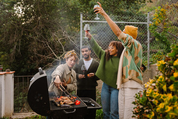 Several multiracial people are standing near the BBQ grill and smiling, some of them raising their...
