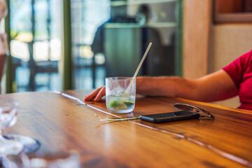 Cold drink with ice, lime, and straw on wooden table near smartphone and sunglasses, with man resting arm nearby. Mexico.