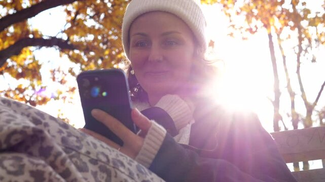 Autumn sunlight filtering through vibrant leaves, woman in white beanie and black coat sitting on park bench, scrolling smartphone. concept of information consumption from Internet