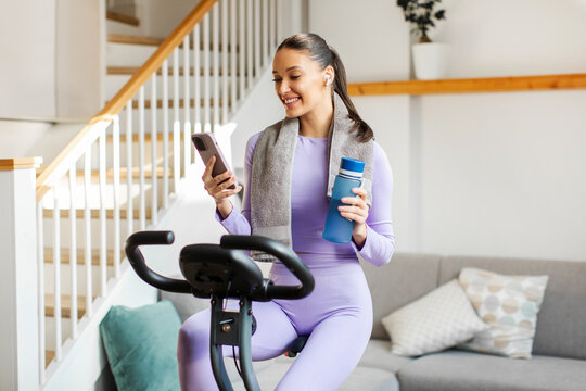 Smiling fitness lady using cellphone and holding bottle of water while training on exercise bike at home in light room