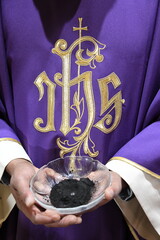 Ein römisch-katholischer Priester hält am Aschermittwoch eine Schale mit scharzer Asche. Der lilafarbene Talar, hat die Aufschrift: IHS. Iesus Hominum Salvator. Jesus, Erlöser der Menschheit 