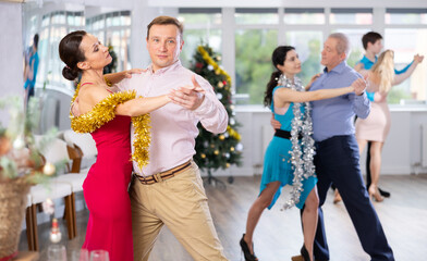 Elegant adult couple, woman and man adorned with sparkling New Year decorations enjoying slow foxtrot at office celebration..