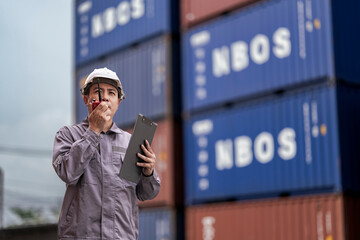 A logistics supervisor in a hard hat and uniform communicates via radio while holding a clipboard...
