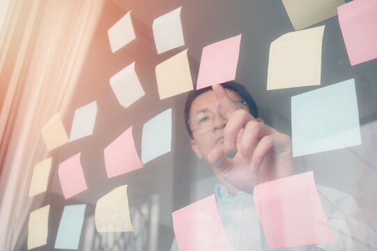 A man in casual attire, standing at an office notice mirror wall board with colorful post-it notes and complex flow charts, focusing on creating creative designs for marketing material