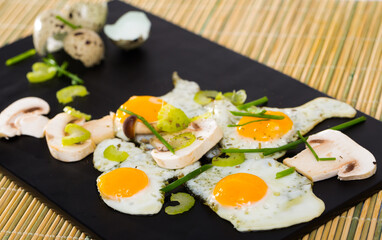 Delicious broken quail eggs with green onion and sliced champignons on black serving board