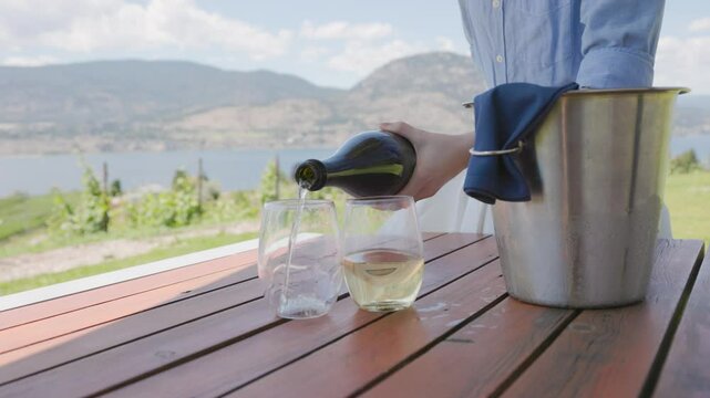 Woman's hand pouring bottle of sparkling wine champagne in glasses on patio overlooking beautiful lake country vista, Okanagan, wine, vineyard. 4K 24FPS