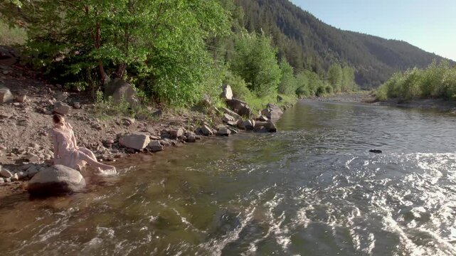 Attractive young woman in a summer dress relaxing beside a mountain river on a sunny day, nature, stream, paradise. 4K 24FPS