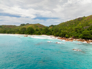 Fototapeta premium A calm coastline with bright turquoise waves meeting a sandy beach, surrounded by lush greenery. Anse Lazio, Seychelles.