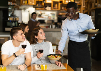 African man waiter serves cafe establishments couple visitor, puts food on table.Restaurant employee presents customer with treat from chef, communicates nicely and wishes them pleasant appetite.