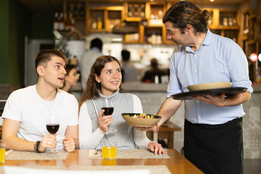 In cozy restaurant hall, man waiter brought couple visitor order, puts traditional Italian salad of leafy greens, vegetables and cheese on table. Waiter cordially serves customers - Powered by Adobe