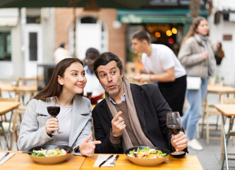 Married couple eating delicious food in restaurant, while sitting on veranda in cool weather. Husband and wife drinking wine and chatting in a European restaurant