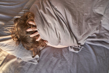 Obraz premium Female caucasian adult wrapped in grey blanket lying in bed with hands on head in relaxed morning mood.