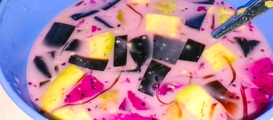 A bowl of fresh fruit soup containing dragon fruit, pineapple, grass jelly, and milk, creating a...