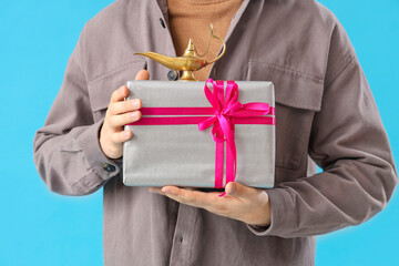 Young Muslim man with gift box and Aladdin lamp on blue background, closeup. Ramadan celebration