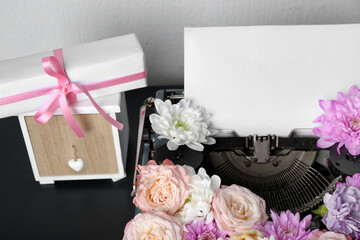 Vintage typewriter with paper sheet, flowers and gift box on black table against white wall
