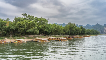 Bamboo tourist rafts are moored in rows by the riverbank. Green vegetation on the shore. Mountains against the sky and clouds. China. Li River. Li Jiang. Guilin