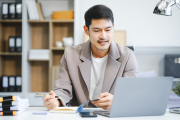 An Asian businessman sits in the office, smiling while using a laptop. He manages business tasks, communicates with clients, analyzes data, and oversees projects to ensure success and productivity