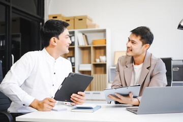 Two Asian businessmen sit in the office using laptops. They manage business tasks, communicate with clients, analyze data, and oversee projects to ensure success and efficiency in their operations