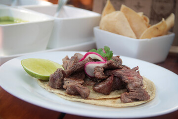Grilled carne asada taco on a soft corn tortilla, topped with radish slices, fresh cilantro, and a lime wedge, served in a tropical beach setting.
