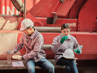 Two industrial workers taking a break at a construction site, wearing safety helmets and protective gear, sitting near heavy machinery, one drinking water and the other taking notes.