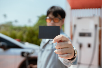 Young man holds a blank fuel card at a gas station on a sunny day, preparing to refuel his car in a rural location