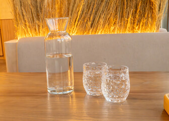 A glass carafe filled with water is accompanied by two intricately designed drinking glasses, set on a smooth wooden table. The ambient lighting and blurred background features dried decorative grass 