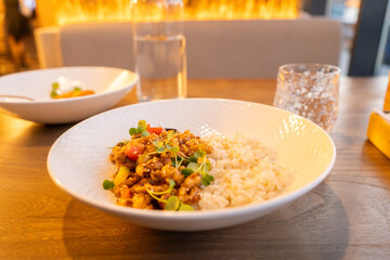 A beautifully plated dish featuring a rice bowl with stir-fried vegetables and garnished with fresh microgreens. The meal is served in a white dish on a wooden table, accompanied by a glass of water