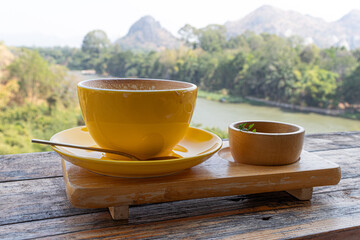 Yellow coffee cup and coffee spoon on wooden table with scenic mountain and river view.