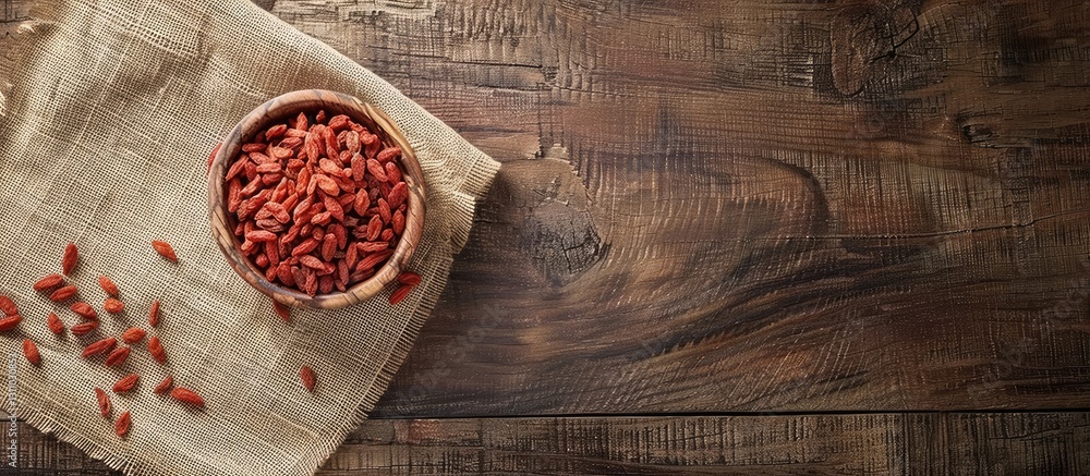 Poster brown wooden background with a rustic cloth; a round bowl filled with red goji berries positioned in