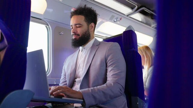 An accomplished African American businessman in a stylish suit is focused on his laptop while comfortably seated on a train, embodying professionalism and high productivity in transit