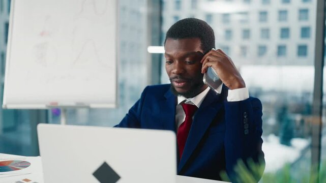 Boss in office, portrait of black man talking by phone and looking at laptop. Nervous African businessman or financial analyst viewing sales diagram and changing strategy of company development