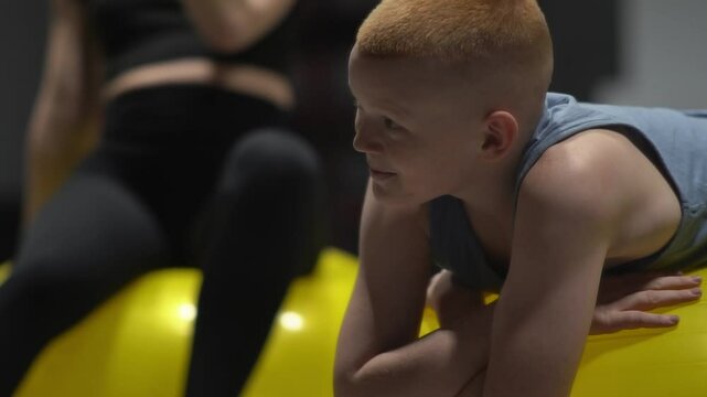 Portrait of red-haired teenager relaxing on fitball and chatting with trainer or mom