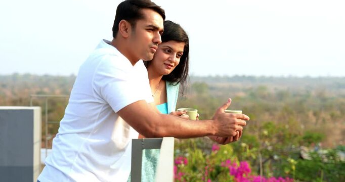 Indian Asian young beautiful happy couple standing on balcony at railing, chatting, laughing, pointing at something far away, enjoying a relaxed morning together drinking coffee outdoors in fresh air
