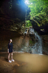 Obraz premium A woman stands near a waterfall falling from a cliff into a mountain river. The gorge. Tourism and recreation