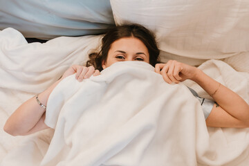 Smiling woman playfully hiding under a soft blanket in bed
