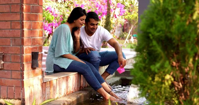 Indian Asian beautiful happy young couple sitting by a water body, playfully dropping a paper boat into the water, laughing, reliving childhood memories, enjoying a joyful, carefree moment outdoors