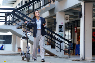Obraz premium Young businesswoman walking with her electric scooter and using a smartphone in urban environment, embracing modern commuting and technology