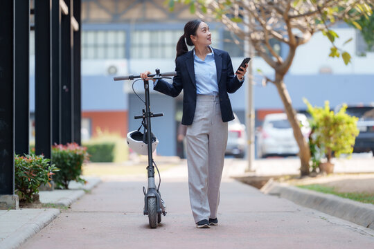 Young businesswoman walking along a city street, using her smartphone while carrying an electric scooter after a busy work commute. Enjoying a modern, eco-friendly lifestyle