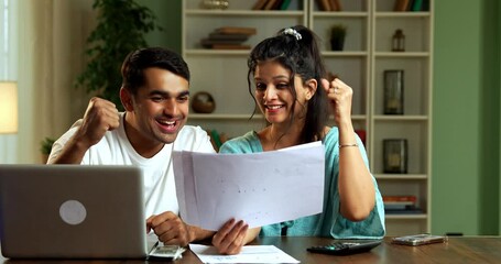 Indian Asian beautiful young couple in late twenties sitting at a desk at home managing finances together, wife looking impressed, happy with husband's financial skills, both celebrating their success
