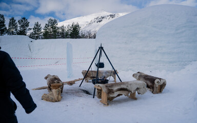 A cozy outdoor seating area, Wooden benches covered in soft reindeer hides encircle a black metal tripod with a hanging pot and a fire pit, perfect for warming up in the Arctic chill. Sami style