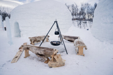 A cozy outdoor seating area, Wooden benches covered in soft reindeer hides encircle a black metal tripod with a hanging pot and a fire pit, perfect for warming up in the Arctic chill. Sami style