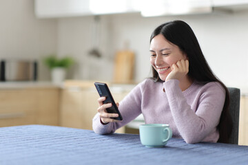 Happy asian woman checking phone content in a kitchen