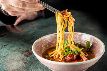 A close-up of a hand using chopsticks to lift steaming noodles from a bowl. The bowl is filled with...