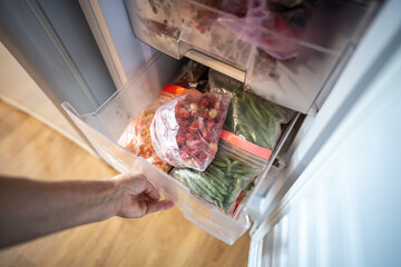 Female hands retrieving frozen raspberries and green beans from fridge box, food in zip bags. Winter supply, fresh berries for desserts, drinks, vegetables for salads, preserving nutrients. Top view