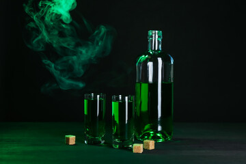 Absinthe in shot glasses, bottle and brown sugar on table against black background with smoke