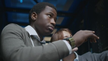 Low angle shot of young confident African American male IT technician analyzing work of hardware with young Caucasian female colleague using laptop, working together in data center - Powered by Adobe